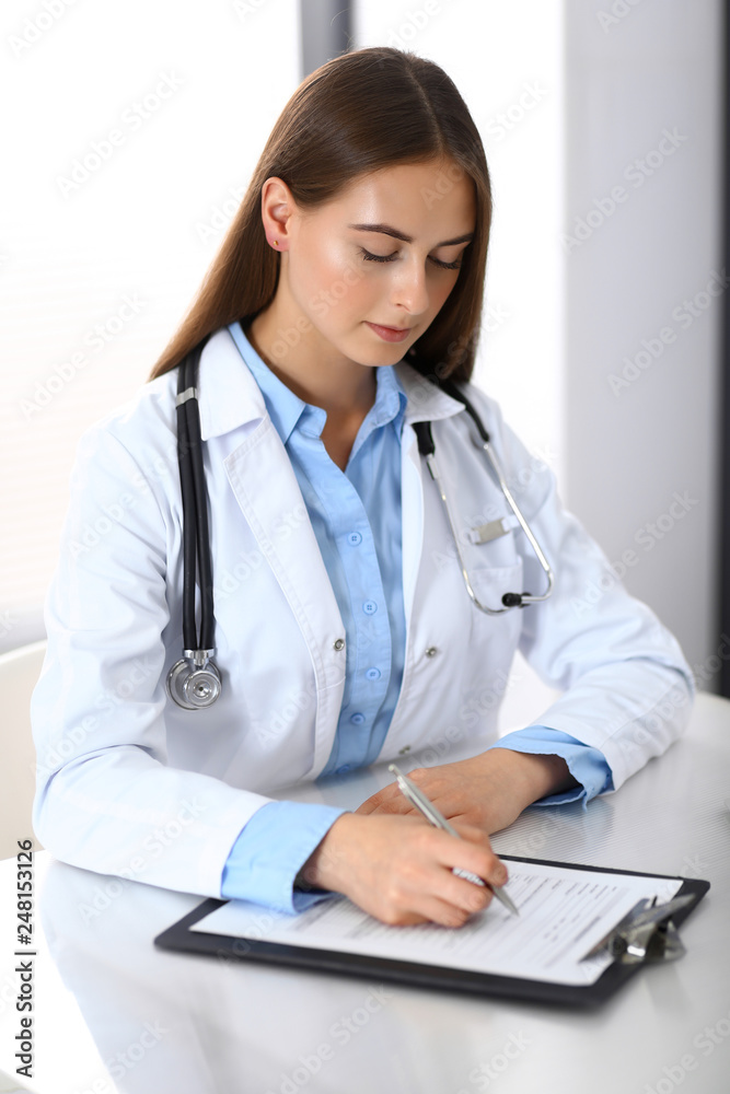 Doctor woman filling up medical form while sitting at the desk in hospital office. Physician at work. Medicine and health care concept Doctor woman filling up medical form while sitting at the desk in hospital office. Physician at work. Medicine and health care concept