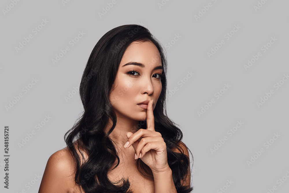 Can you keep a secret? Beautiful young woman looking at camera and keeping finger on lips while standing against grey background Can you keep a secret? Beautiful young woman looking at camera and keeping finger on lips while standing against grey background