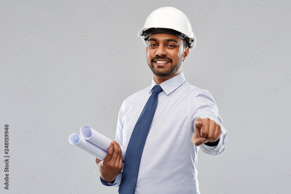 architecture, construction business and building - smiling indian architect or businessman in helmet with blueprints pointing finger to you over grey background architecture, construction business and building - smiling indian architect or businessman in helmet with blueprints pointing finger to you over grey background