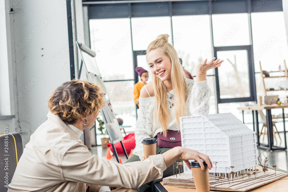 female and male architects with coffee to go looking at each other while working on house model in loft office female and male architects with coffee to go looking at each other while working on house model in loft office