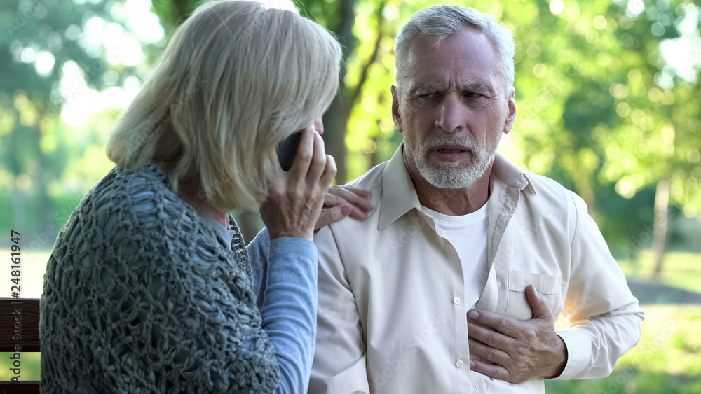 Fototapeta premium Old couple sitting on bench, man suffering arrhythmia, wife calling emergency