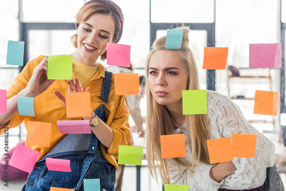 beautiful casual businesswomen looking at colorful sticky notes on glass window in office beautiful casual businesswomen looking at colorful sticky notes on glass window in office