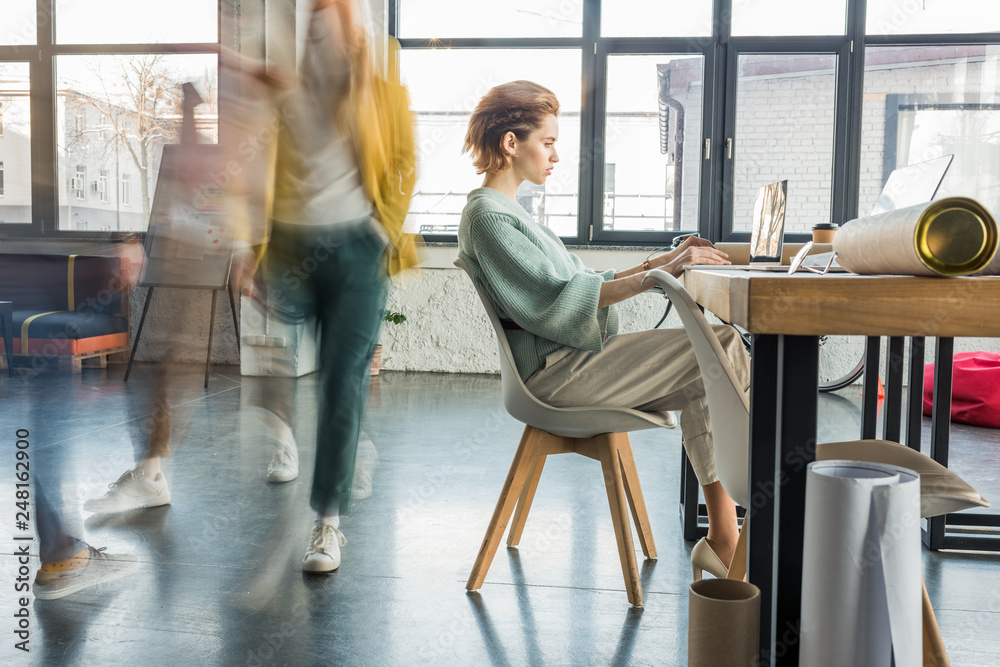 female architect sitting and using laptop at desk in loft office with colleagues in motion blur on background female architect sitting and using laptop at desk in loft office with colleagues in motion blur on background