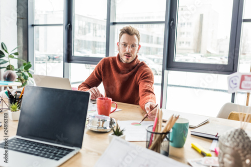 handsome casual businessman sitting at computer desk, looking at camera and working on document in loft office