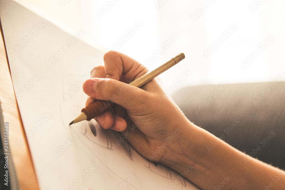 Close up view of a young hand drawing on a white sheet. Kid hold a black wooden pencil and draw something on a warm orange light at home. Children writing on a paper. Teen drawing freehand a manga. - obrazy, fototapety, plakaty 