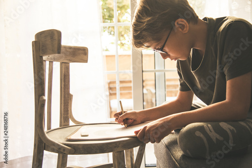 Close up view of a young boy drawing on a white sheet. Kid hold a pencil and draw something, warm orange light at home Children drawing freehand on a paper in front of a window using a chair as a desk photo