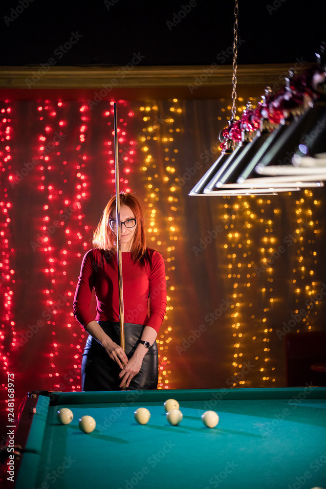 Billiard club. A woman with in glasses with red hair and nice figure standing by the table holding a cue