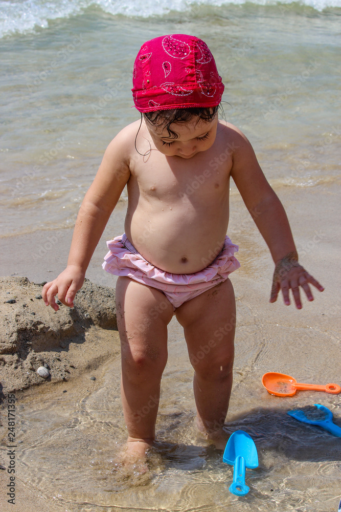 A cute and sweet little girl plays with water and sand on the seashore. Child with pink swimsuit and red bandana, plays with a plastic scoop. Sea foam and waves. A cute and sweet little girl plays with water and sand on the seashore. Child with pink swimsuit and red bandana, plays with a plastic scoop. Sea foam and waves.