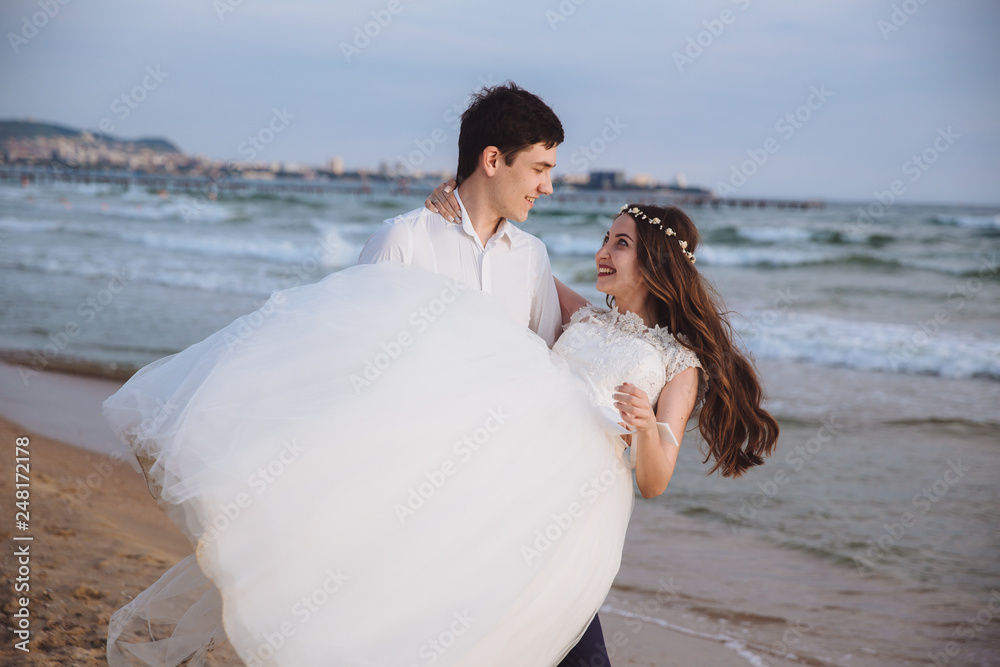 Groom carries bride in his arms on ocean beach. Just married happy couple have fun at wedding day Groom carries bride in his arms on ocean beach. Just married happy couple have fun at wedding day