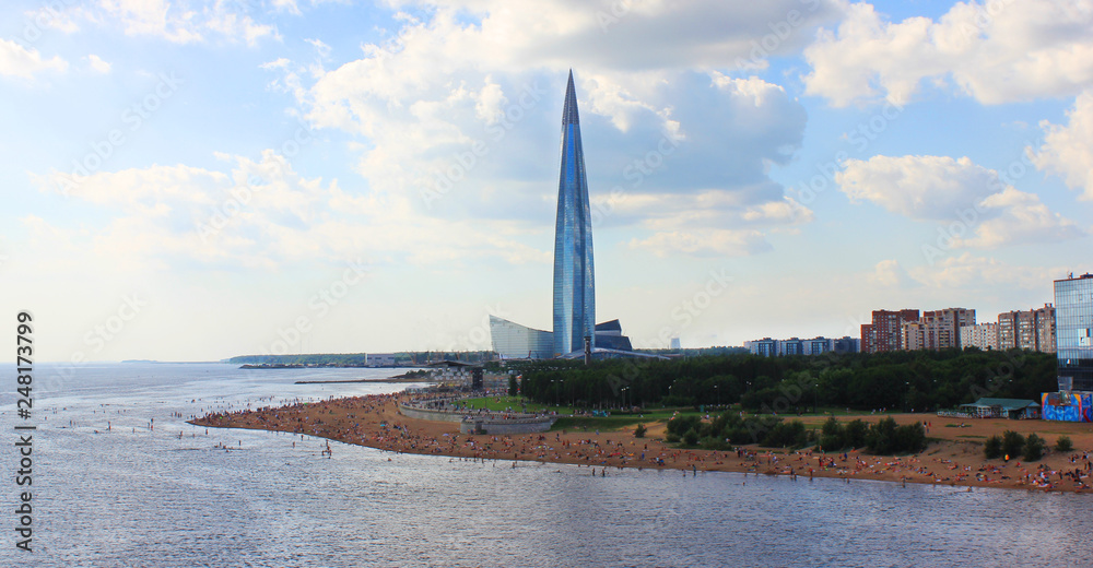 Modern Skyscraper Architecture View with Public City Beach and People Relaxing on Summer Day. Urban Tall Building in Downtown Area Facing the Park and Sandy Beach in Saint Petersburg, Russia