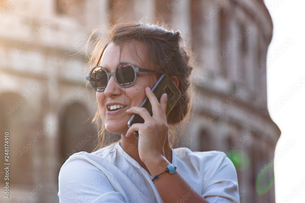 Beautiful young woman with smartphone in fashion dress standing in front of colosseum in Rome at sunset. Talking on phone Beautiful young woman with smartphone in fashion dress standing in front of colosseum in Rome at sunset. Talking on phone