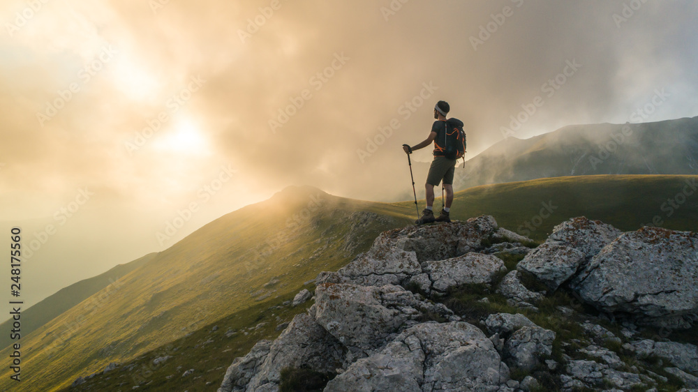 Young hiker man with backpack and walking poles, standing on peak of a mountain looking at sunset in cloudy sky. Green field and rocks. Abruzzo, Italy.