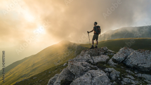Young hiker man with backpack and walking poles, standing on peak of a mountain looking at sunset in cloudy sky. Green field and rocks. Abruzzo, Italy. photo
