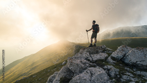 Young hiker man with backpack and walking poles, standing on peak of a mountain looking at sunset in cloudy sky. Green field and rocks. Abruzzo, Italy. photo