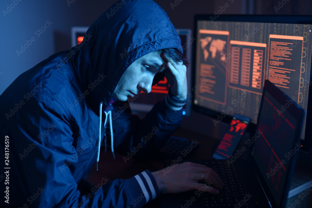 Hacker in a hood thinking about the problem of hacking or malware infection on the background of screens with codes
