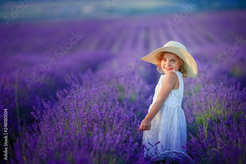 Cute curly young girl standing on a lavender field in white dress and hat with cute face and nice hair with lavender bouquet and smiling.