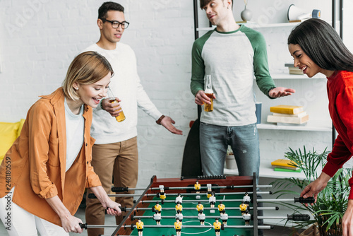 smiling multicultural girls playing table football while men talking and drinking beer