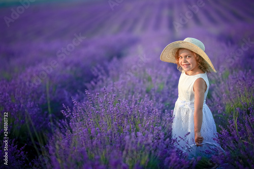 Cute curly young girl standing on a lavender field in white dress and hat with cute face and nice hair with lavender bouquet and smiling.