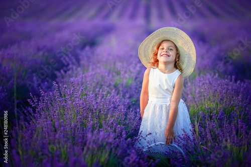 Fototapeta Naklejka Na Ścianę i Meble -  Cute curly young girl standing on a lavender field in white dress and hat with cute face and nice hair with lavender bouquet and smiling.
