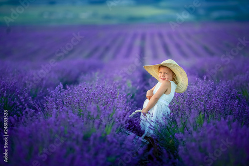 Cute curly young girl standing on a lavender field in white dress and hat with cute face and nice hair with lavender bouquet and smiling.