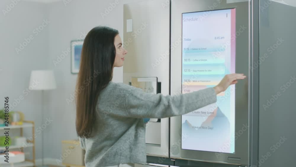 Beautiful Young Girl Walks Over to a Refrigerator While Drinking Her Morning Coffee. She is Checking the Weather Forecast and a To Do List on a Smart Fridge at Home. Kitchen is Bright and Cozy.