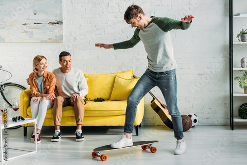 multicultural friends sitting on yellow sofa and looking at friend riding longboard