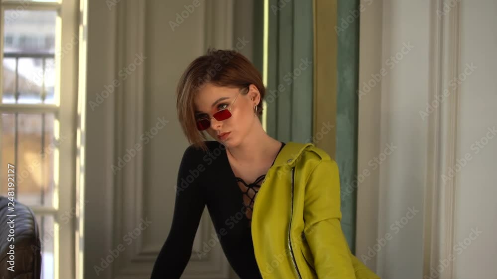 A brunette woman with a short haircut quads and sunglasses posing. She's wearing a black bodysuit and a light green leather jacket. A brunette woman with a short haircut quads and sunglasses posing. She's wearing a black bodysuit and a light green leather jacket.