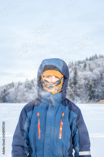 Arctic outdoor winter portrait of a child boy in winter clothing with balaclava against frozen landscape background.