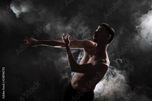A young male ballet dancer with black leggings and a naked torso performs dance moves against a gray grunge background, with a light of lights and smoke. © spaskov