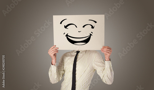 Casual person holding a paper in front of his face with drawn emoticon face 
