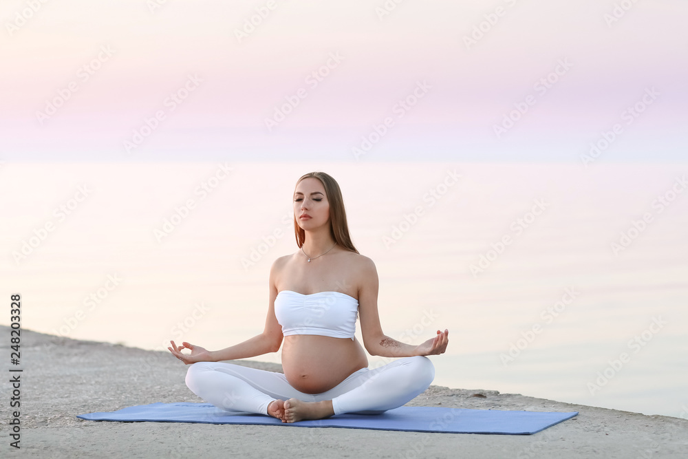 Young pregnant woman in white dress sitting on the beach near blue sea and breathing. Summer vacation during pregnancy, happy motherhood concept, close up on hands