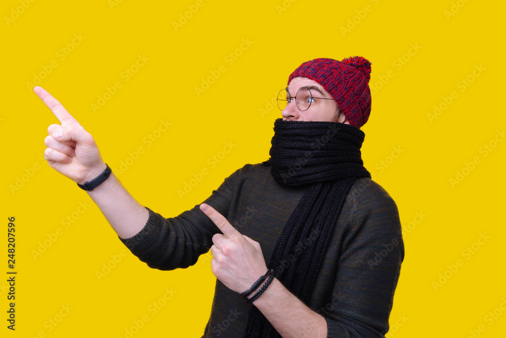 Young man in gray sweater, black scarf and red hat pointing index fingers away. Cold season concept Young man in gray sweater, black scarf and red hat pointing index fingers away. Cold season concept