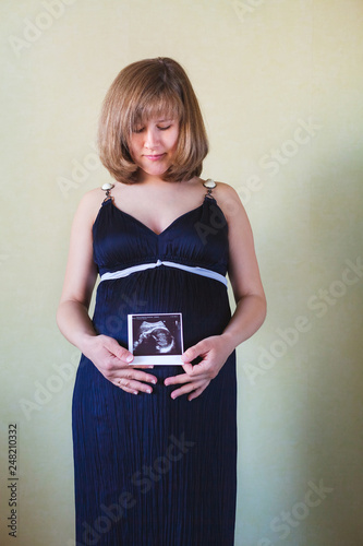 Pregnant woman wearing deep blue dress standing with ultrasound shot above her belly over green walll