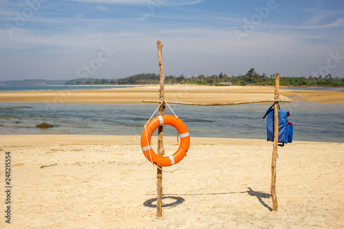 orange lifebuoy ring and blue life vest on a bamboo stand on a sandy beach against the backdrop of the sea bay and green jungle