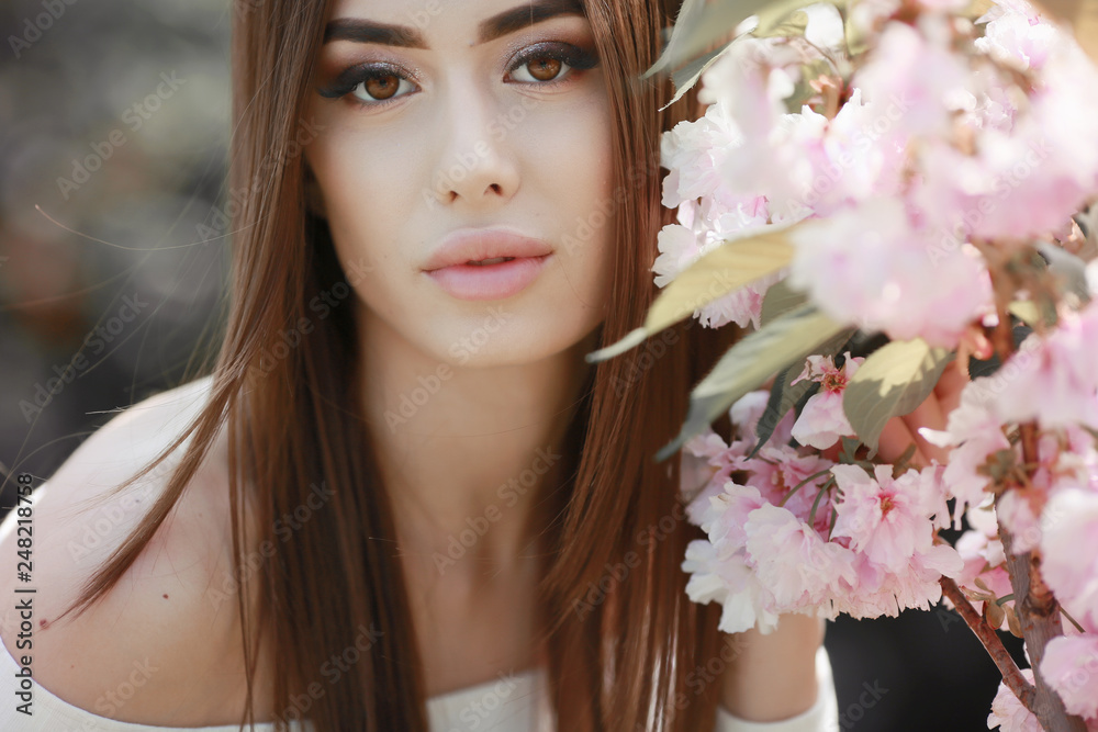 The beautiful girl with a romantic hairstyle and a professional make-up enjoys a smell of pink colors in a garden. The girl dreams. A portrait of the beautiful girl model in the spring in the park.