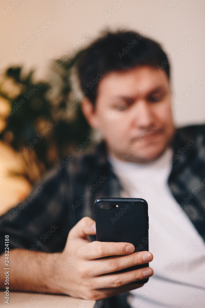 Close up detail of a man's hand holding a smartphone, browsing the internet while sitting in a personal working space. Focus on the device. Close up detail of a man's hand holding a smartphone, browsing the internet while sitting in a personal working space. Focus on the device.