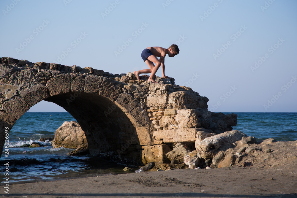 Fototapeta premium Little boy plays on a ruined old stone bridge next to a beach.