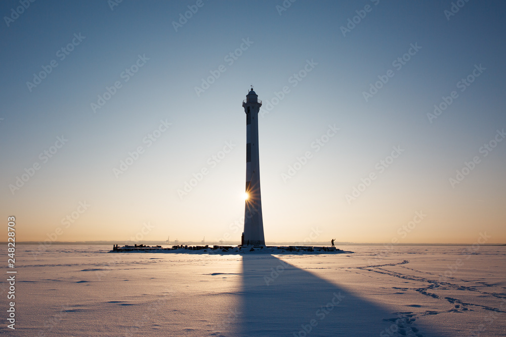 Lighthouse on a frozen and snow covered Gulf of Finland in frosty weather, sun is shining behind the lighthouse, silhouette of man near, copy space. Winter sunshine concept