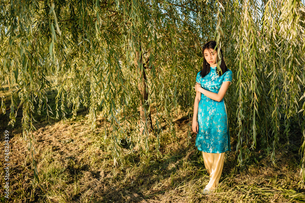 Portrait of beautiful Asian Chinese brunette in blue national dress outdoors, young Vietnamese woman in traditional clothes, the girl is resting in park Portrait of beautiful Asian Chinese brunette in blue national dress outdoors, young Vietnamese woman in traditional clothes, the girl is resting in park