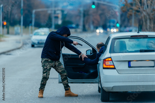 The car thief is pulling the car owner out of his car and trying to get the car while pointing a loaded gun at the drivers head..