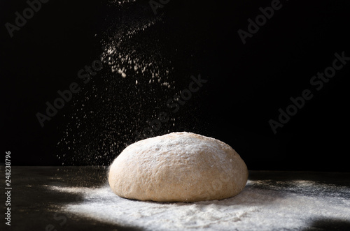 Flour pours on fresh homemade dough. Dough with spilled flour on a black background. Yeast dough for bread, rolls, pizza or pie. Copy space.