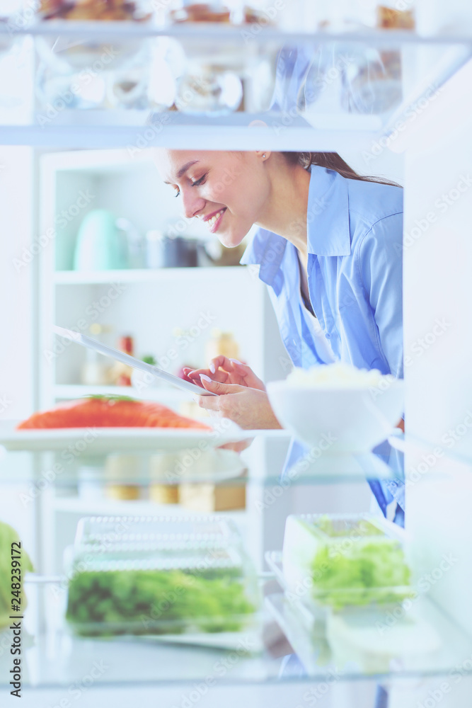 Portrait of female standing near open fridge full of healthy food, vegetables and fruits.