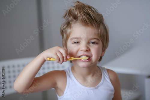 Close up portrait of cute, adorable, toddler boy wearing denim overalls, long T-shirt, sitting on the floor, brushing his teeth with a toothbrush