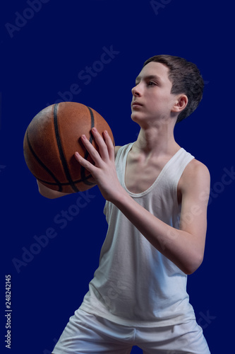 Teen basketball player. The guy in the white T-shirt holds the ball in his hands and is preparing to throw it. Template for the design of a sports poster or news about competitions. Isolated on blue. © romsvetnik