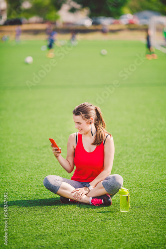Theme sport health. beautiful young girl sitting resting on green grass. lawn stadium using technodogies. In handphone in ear headphones listens music, summer sportswear watch heart rate monitor