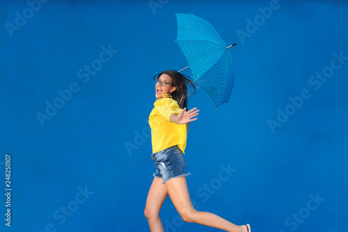 Cute teenage girl with brown hair and eyeglasses wearing yellow blouse and denim shorts while running with blue umbrella with black dots. Blue background.