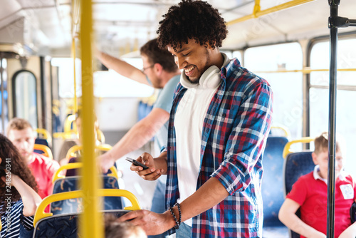 Young smiling African American man using smart phone and standing while riding in the city bus.