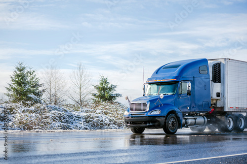 Big rig blue semi truck tractor transporting commercial cargo in refrigerator semi trailer going on the wet road with melting snow with winter snowy trees on the side