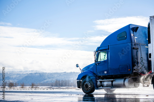 Blue big rig long haul semi truck with reefer semi trailer running on winter wet road with melting snow and water dust photo