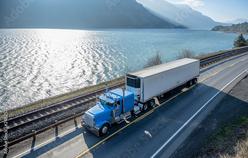 Blue classic American bonnet big rig semi truck trancporting cargo in refrigerated semi trailer moving on the road along the river in Columbia River Gorge area